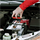 A hand places a red device with symbols into a holder next to a grey car battery in an engine bay.