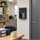 Black wall-mounted deposit safe with a drop slot, lower door open with a key in the lock, in an office environment.