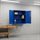 Blue metal wall cabinet with two open doors and three shelves, showing boxes and binders inside, in a workshop environment.